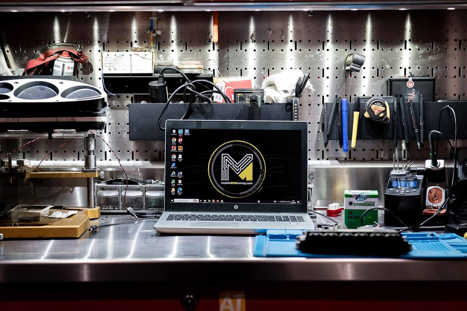 A professional automotive workstation with a laptop displaying a tuning company's MET logo on its screen. The laptop is set on a metallic workbench surrounded by various car tuning tools and equipment neatly arranged on a pegboard in the background, indicating a setting for vehicle ECU mapping services.