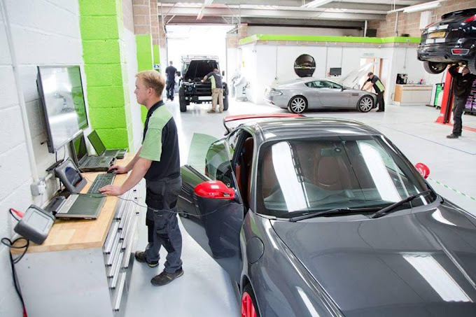 A mechanic works on a laptop beside a grey sports car with red mirrors in a garage. The car's door is open, and other cars and technicians are visible in the background.