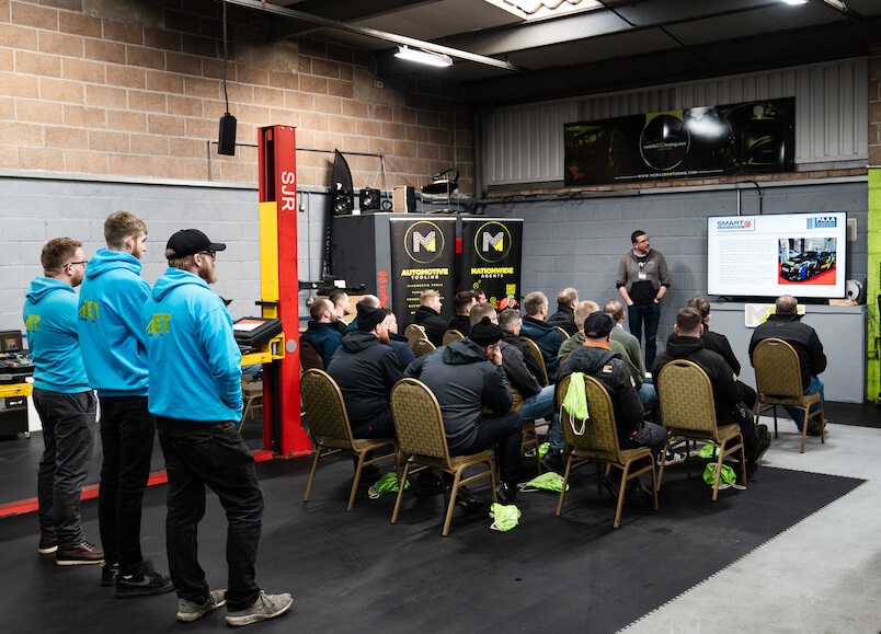 A group of attendees in a garage, listening to a presenter at the Mobile Eco Tuning open day. The presenter is speaking next to a large screen displaying technical content. The audience, mostly wearing matching turquoise jackets, is seated or standing around in an attentive posture.